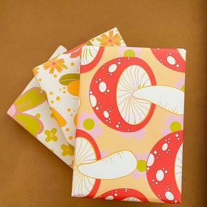 May include: Three sheets of wrapping paper with a mushroom and floral pattern. The top sheet is a light orange with red and white mushrooms. The middle sheet is white with orange and yellow flowers. The bottom sheet is green with pink and yellow flowers.