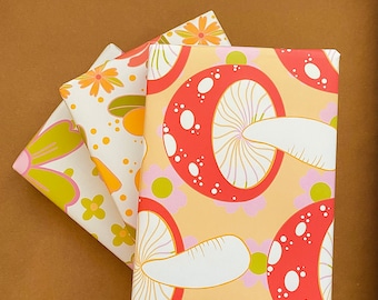 May include: Three sheets of wrapping paper with a mushroom and floral pattern. The top sheet is a light orange with red and white mushrooms. The middle sheet is white with orange and yellow flowers. The bottom sheet is green with pink and yellow flowers.