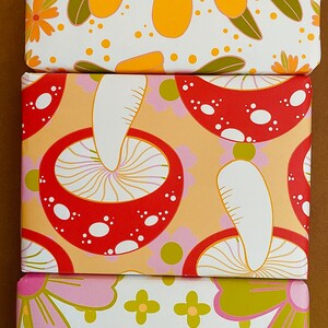 May include: Three different wrapping paper designs featuring a retro 70s style. The first design has yellow and orange flowers on a white background. The second design has red and white mushrooms on a light orange background. The third design has pink and green flowers on a white background.