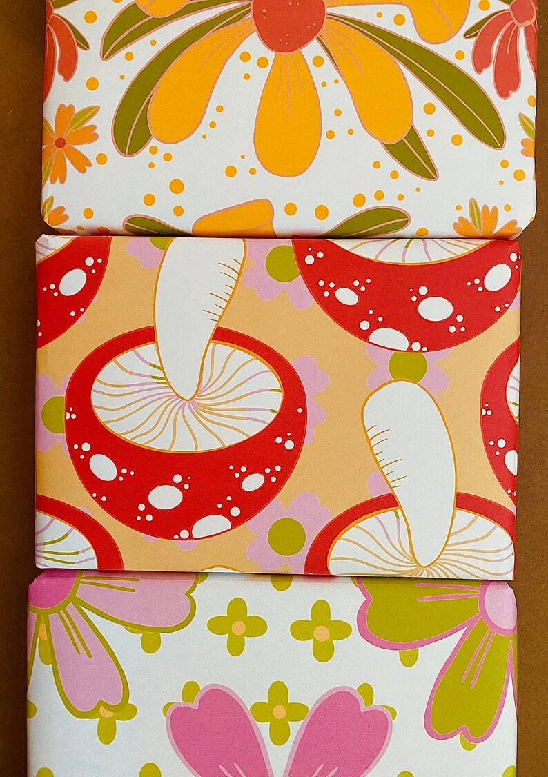 May include: Three different wrapping paper designs featuring a retro 70s style. The first design has yellow and orange flowers on a white background. The second design has red and white mushrooms on a light orange background. The third design has pink and green flowers on a white background.