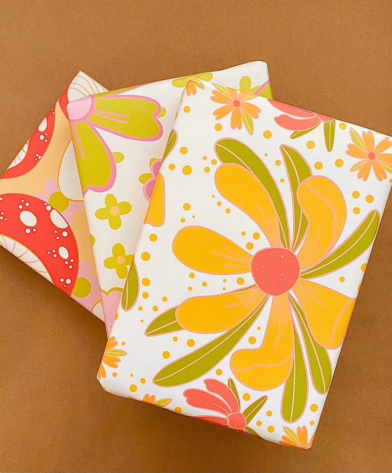 May include: Three sheets of wrapping paper with a retro floral design. The paper features large yellow flowers with pink centers and green leaves on a white background. One sheet has a mushroom design with red and orange mushrooms on a yellow background. The other sheet has a pink and green floral design on a white background.