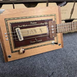 May include: A handmade cigar box guitar with a wooden body and a fretboard. The guitar features the text "JAVA" and "Drew Estate" on the front. It has metal hardware and strings, with a toggle switch and tuning pegs.