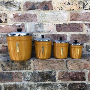 May include: A set of four vintage kitchen canisters in a warm yellow hue, with silver rims and black knobs. The canisters are labelled with text and have a wood-grain appearance, arranged against a brick wall.