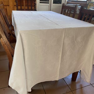 May include: A white rectangular tablecloth with a delicate floral design covers a wooden table. Wooden chairs surround the table. The tablecloth hangs over the edges, forming gentle folds. The room has a rustic atmosphere.