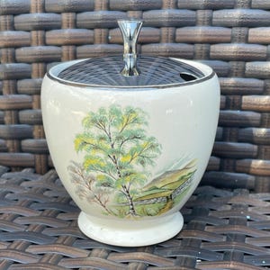 May include: A vintage white ceramic sugar bowl with a lid and a silver handle. The bowl features a painted landscape scene with trees and a fence. The lid has a black and silver striped pattern.