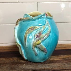 May include: A turquoise ceramic vase with a leaf design. The vase has a rounded shape with a textured surface and a light yellow interior. The leaf design features yellow and maroon accents.