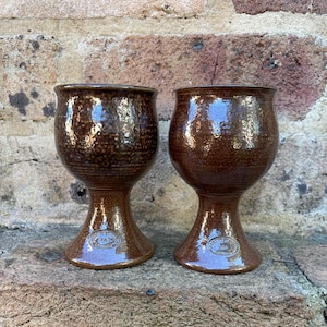 May include: Two brown ceramic goblets with a glossy, speckled glaze finish. Each goblet features a wide, rounded bowl, a tapered stem, and a flared base. A small, circular maker's mark is embossed on the lower portion of each stem.