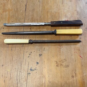 May include: Three antique sharpening tools. The top tool has a wooden handle and a metal blade. The middle tool has a cream-coloured handle and a metal blade. The bottom tool has a cream-coloured handle and a metal blade.