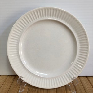 May include: A white ceramic plate with a ridged rim. The plate is round and has a slightly textured surface. The plate is displayed on a clear plastic stand, suggesting it is for decorative or display purposes.