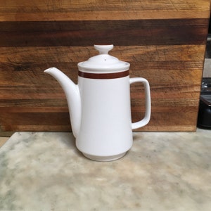 May include: White ceramic coffee pot with a brown stripe around the top. The pot has a lid and a handle.