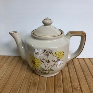 May include: A beige ceramic teapot with a brown handle and lid. The teapot is decorated with a floral pattern of yellow and white flowers.