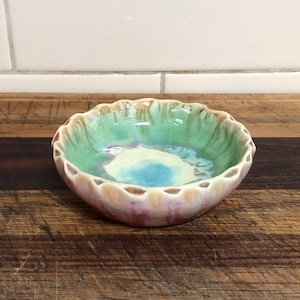 May include: A small, handmade ceramic bowl with a scalloped rim. The interior features a blend of turquoise, light green, and blue hues, while the exterior displays shades of pink and peach. The bowl is approximately 15 cm in diameter.