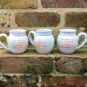 May include: Three white ceramic mugs with a light blue rim and a pink floral design. The mugs have a rounded shape and a handle.
