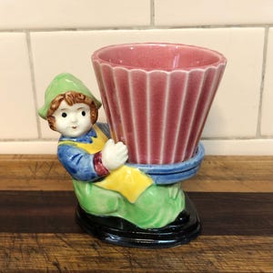 May include: A vintage ceramic planter depicting a seated figure holding a ribbed, pink vase. The figure is dressed in a green hat, blue shirt, yellow overalls, and green trousers. The base is black, on a wooden surface.