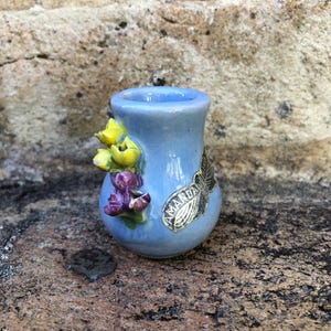 May include: A small, light blue ceramic vase with a floral design. The vase has a yellow and purple flower design on one side and a black and white fish design on the other side. The vase is sitting on a rock.