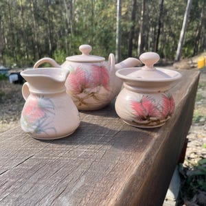 May include: A pale pink ceramic tea set comprising a teapot, creamer jug, and sugar bowl. Each piece features a pink and green floral design. The teapot has a lid and handle, the creamer a handle and spout, and the sugar bowl a lid.