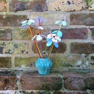 May include: A turquoise ceramic vase with a floral design holds a bouquet of colorful paper pinwheels. The pinwheels are made of blue, green, red, and white paper and have a whimsical, handmade look.