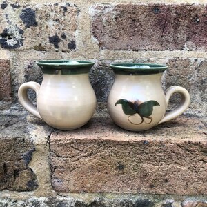 May include: Two ceramic mugs with a cream-coloured body and a dark green rim. One mug features a green leaf design. The mugs have a rounded shape with a handle. The mugs are sitting on a brick surface.