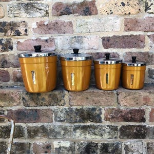 May include: A collection of four vintage kitchen canisters in different sizes. They are a golden yellow with silver lids and black knobs. Each canister has a rectangular, metallic accent. The canisters are displayed on a brick ledge, against a brick wall.