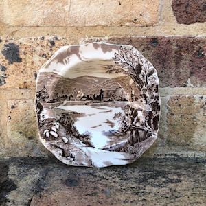 May include: A brown and white ceramic bowl with a scenic landscape design. The bowl features a brown and white landscape with a river, trees, and a farmhouse. The bowl is in a rustic style.