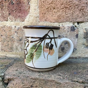 May include: A ceramic mug with a cream-coloured base and a dark brown rim. The exterior features a hand-painted botanical design with green leaves and light orange berries, accented by dark brown branches and horizontal bands around the top and bottom.