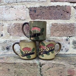 May include: Three ceramic mugs with a speckled tan and olive green glaze. Each mug features a hand-painted design of a stylised red mushroom cap on a dark brown branch with green leaves. The mugs have a cylindrical shape and a curved handle.
