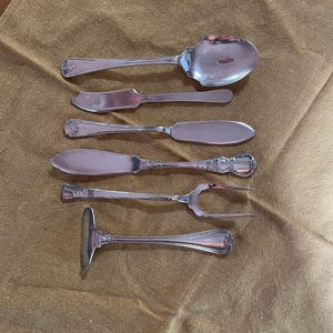 May include: An assortment of antique silver-colored silverware, including a spoon, butter knives, a fork, and a serving utensil. The silverware is arranged on a brown cloth, showcasing the intricate details and designs of each piece.