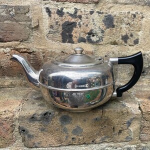 May include: A polished silver teapot with a black handle and a lid. The teapot is round with a spout and sits on a brick surface. The teapot reflects the surrounding environment. The handle is black.