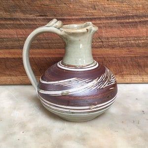 May include: A brown and white ceramic jug with a distinctive handle. The jug has a round body with a wide mouth and a narrow neck. The jug is decorated with brown and white stripes and a white glaze.