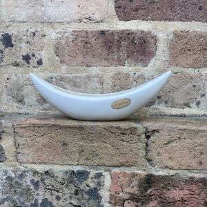 May include: A smooth, off-white ceramic planter shaped like a crescent moon or a boat. The vessel features a curved, elongated silhouette with pointed ends and a small, oval-shaped gold-toned label on the front surface with the text Raynham.