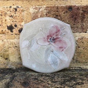 May include: A ceramic dish with a raised floral design. The dish is light grey with a pink flower and green leaves.