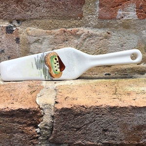 May include: White ceramic cake server with a painted design of a slice of cake with chocolate and green frosting. The server has a handle with a hole in it.