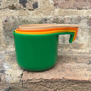 May include: A set of four brightly coloured measuring cups nested together. The largest cup is green, with orange, yellow, and beige cups inside. The cups are plastic and have a handle.