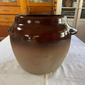 May include: A large, antique brown stoneware crock with a two-tone finish. The upper section is a glossy, dark brown, while the lower half has a textured, lighter brown appearance. It has two small handles on either side.