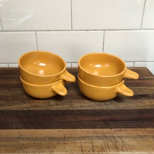 May include: Four mustard-yellow ceramic soup bowls, stacked in pairs, each with a handle. The bowls have a rounded shape and a small handle on one side. They are on a wooden chopping board.