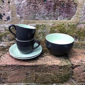 May include: Three stacked dark grey teacups with a light green interior and a matching saucer with a light green and white floral pattern. A dark grey bowl with a light green interior is also shown.