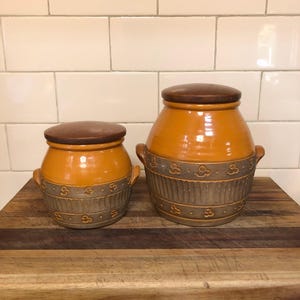 May include: Two ceramic canisters with wooden lids. The canisters are brown and orange with a decorative pattern. The larger canister is taller than the smaller canister.