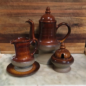May include: A brown ceramic teapot, creamer and sugar bowl with lids. The teapot has a brown glaze with a speckled finish. The creamer and sugar bowl have a similar glaze and finish.