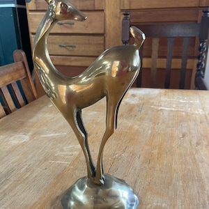 May include: A polished brass figurine of a standing deer with a smooth, reflective surface. The deer is depicted with its head turned to the side, featuring slender legs and a short tail, all mounted on a rounded, uneven brass base.