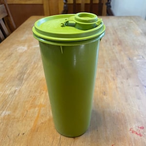 May include: A green plastic tumbler with a matching lid. The lid features a flip-top opening and a raised drinking spout. The cylindrical tumbler appears empty. The design is simple and functional, suitable for everyday use.