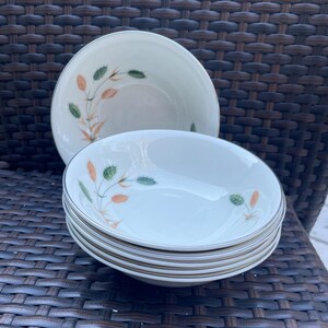 May include: A collection of white ceramic bowls featuring a floral pattern in green and orange, with a silver rim. Several bowls are stacked, with one separate in the background. The bowls are displayed on a brown woven surface.