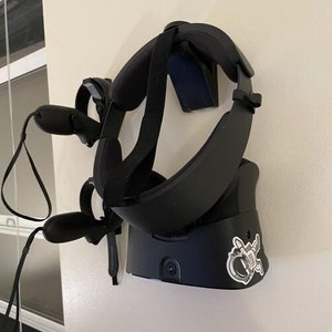 May include: A black virtual reality headset with two controllers hanging on a white wall. The headset has a black strap and a sticker on the front that says "X".