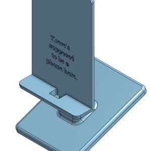 May include: A light blue phone stand with a rectangular base and a vertical stand. The stand has a cutout for a phone and the text "There's supposed to be a phone here." is printed on the vertical stand.