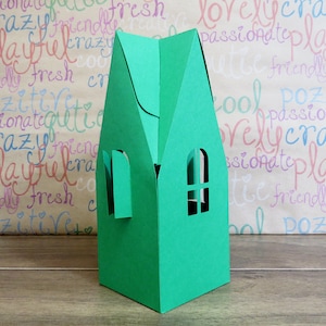 May include: A green paper house-shaped gift box with two window cutouts. The box has a folded roof and is set against a background with colorful handwritten words. The box is on a wooden surface.