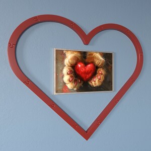 May include: A red heart held in the paws of a cat, framed in a red heart-shaped frame against a blue wall.