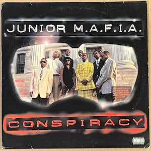 Pode incluir: Uma capa de álbum preta e vermelha com o título "JUNIOR M.A.F.I.A. CONSPIRACY" em letras brancas. A capa apresenta um grupo de homens em frente a um edifício com colunas.