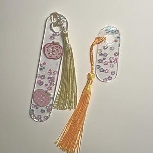 May include: Two clear resin bookmarks with star and glitter accents. One bookmark features a pink apple design, the other has a star-shaped cutout. Both have colorful tassels, one gold and one orange, attached at the top.