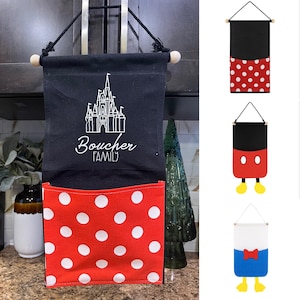 Op de afbeelding: Een wandorganizer van zwarte stof met een rood vak met witte stippen. Het zwarte gedeelte toont een witte lijntekening van een kasteel boven de tekst Boucher FAMILY. Het hangt aan een houten stok met een zwart touw.