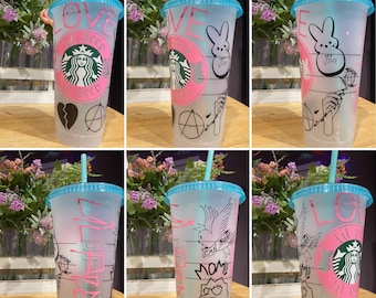 May include: Six clear plastic tumblers with blue lids and straws. Each tumbler has a different design featuring black and pink graphics. The designs include a broken heart, an anarchy symbol, a bunny rabbit, a bird, a cartoon character, and a Starbucks logo.