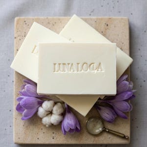 Puede incluir: Tres pastillas de jabón rectangulares de color blanco roto apiladas sobre una baldosa de piedra cuadrada. Cada pastilla lleva grabado el texto LUNALOCA. El conjunto está decorado con flores de azafrán moradas, algodón blanco y una lupa de latón.